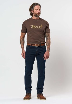 Moški z tatuji, ki nosi rjavo majico z grafiko, temno modre kavbojke, rjav pas in rjave usnjene škornje, stoji na belem ozadju.