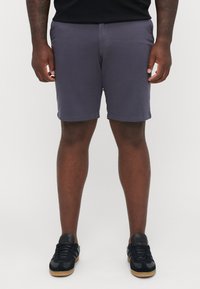 Mørk lilla shorts med en glatt tekstur, sidelommer og et avslappet design, kombinert med svarte joggesko med gummisåler.