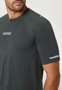 Trumpa rankovė tamsiai pilka sportinė marškinė, pagaminti iš švelnios medžiagos, su balta "GUESS" logotipu ant krūtinės ir pažymėjimu ant rankovės.