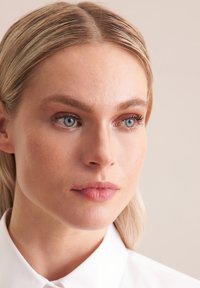 Zbliżenie na osobę o jasnej karnacji, prostych blond włosach i niebieskich oczach, noszącą białą koszulę z kołnierzykiem, z naturalnym makijażem i gładką strukturą skóry.