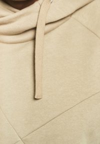 Felpa beige con un tessuto texturizzato, dotata di cappuccio con cordino e cuciture diagonali sul davanti. Design morbido e casual.