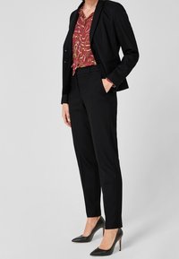 Žena oblékající černý elegantní oblek s vínově červenou vzorovanou halenkou a černými lodičkami, stojící s jednou rukou v kapse.