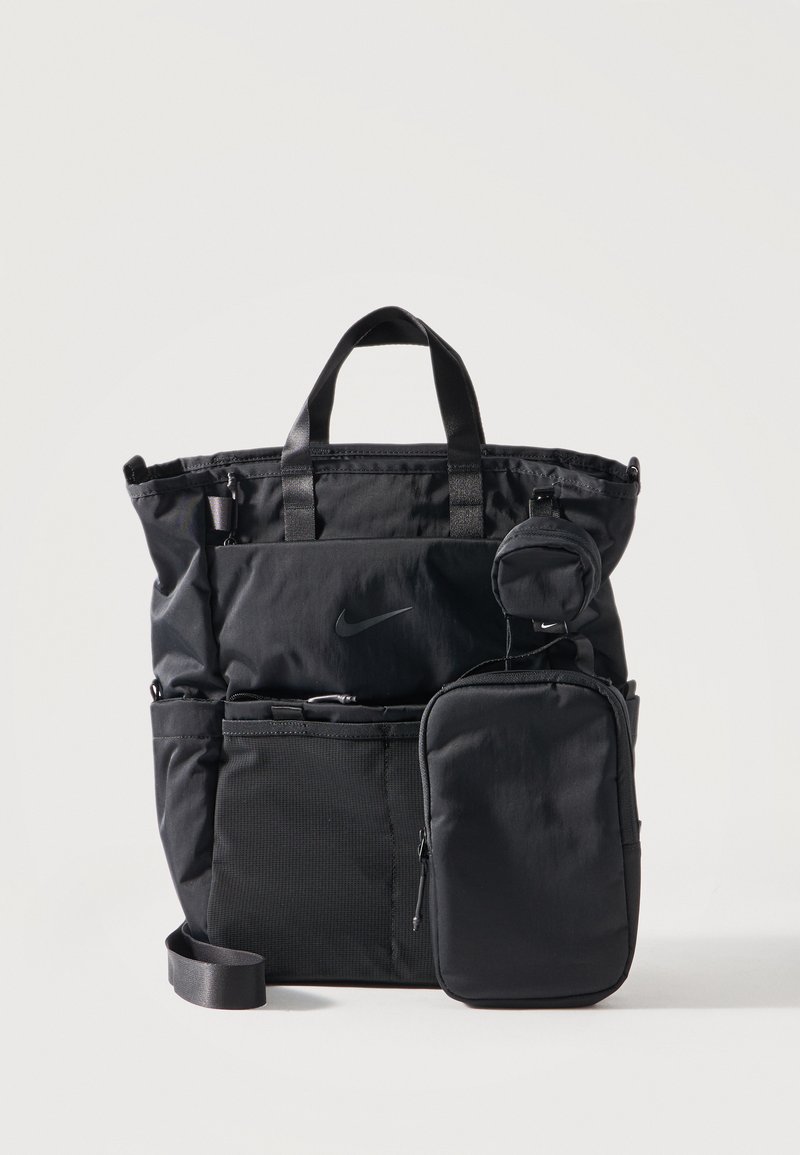 Μαύρη τσάντα Tote Nike με διπλές λαβές, εξωτερική θήκη με φερμουάρ, μικρή στρογγυλή θήκη προσαρτημένη και ιμάντα ώμου σε ανοιχτό φόντο.