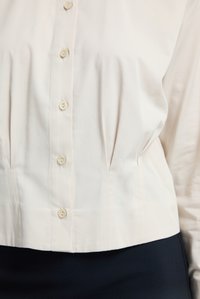 Krämfärgad skjorta med figursydd midja, knappstängning och lätta veck. Mjukt tyg, långa ärmar och kort längd.