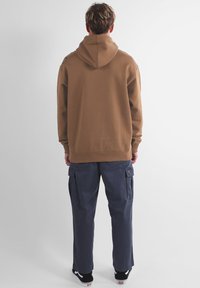 Sweat-shirt à capuche marron de coupe ample ; porté avec un pantalon cargo bleu foncé comprenant des poches latérales ; style décontracté de rue.