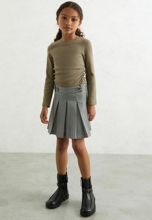 Olivenes Langarmshirt kombiniert mit einem grauen karierten Faltenrock mit Metallknopf-Akzenten; schwarze Ankle-Boots mit robusten Sohlen.