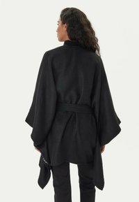 Cardigan nero avvolgente realizzato in tessuto morbido, caratterizzato da maniche larghe, una vita con cintura e un taglio fluido che si raccoglie sui fianchi.
