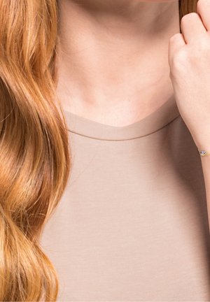 Longs cheveux rouges tombant sur le col d'une chemise beige, avec une main touchant les cheveux et portant un bracelet délicat orné d'une petite pierre violette.