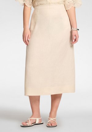 Beiger Midi-Rock getragen mit einer Spitzenbluse, kombiniert mit weißen flachen Sandalen mit Riemchen und einem weißen Armband am rechten Handgelenk.