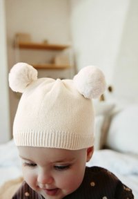 Cappello per neonati in maglia di colore crema chiaro con due pompon morbidi in cima, caratterizzato da un orlo a coste e una texture soffice.