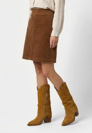 Kobieta ubrana w brązową sztruksową spódnicę za kolano, beżową koszulę z długim rękawem oraz beżowe zamszowe buty za kostkę z szerokim obcasem i bocznymi zamkami.