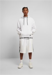 Hvit hettegenser med snor, med en logo-bånd nederst. Kombinert med hvite shorts, også prydet med svarte logo-aksenter. Hvite sneakers.