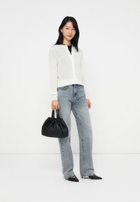 Cardigan léger et blanc à boutons, top noir, jeans évasés bleus, tenant un sac à main noir froncé, et portant des ballerines noires à bouts pointus.