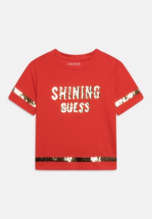 Chemise rouge à manches courtes avec des rayures de sequins dorés sur les manches et le bas, texte scintillant "SHINING GUESS" en sequins dorés sur le devant.