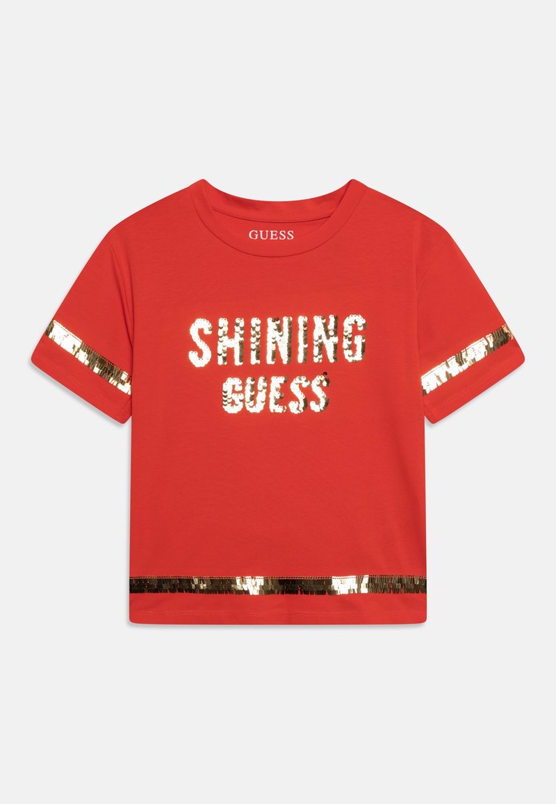 Chemise rouge à manches courtes avec des rayures de sequins dorés sur les manches et le bas, texte scintillant "SHINING GUESS" en sequins dorés sur le devant.