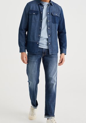 Chaqueta de denim azul oscuro con dos bolsillos en el pecho, combinada con una camiseta de color azul claro y jeans de color azul claro. Unos deportivos blancos casuales completan el look.