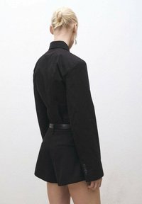 Musta räätälöity bleiseri, jossa on pitkät hihat ja kapea muotoilu, yhdistettynä korkeavyötäröisiin mustiin shortseihin. Silkkinen pinta, minimalistinen tyyli.