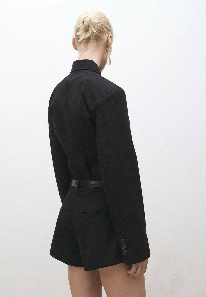 Musta räätälöity bleiseri, jossa on pitkät hihat ja kapea muotoilu, yhdistettynä korkeavyötäröisiin mustiin shortseihin. Silkkinen pinta, minimalistinen tyyli.