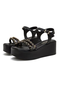 Sandali platform neri con tacco alto, caratterizzati da doppie cinghie alla caviglia e dettagli in catena dorata sulla parte anteriore. Texture in pelle liscia.