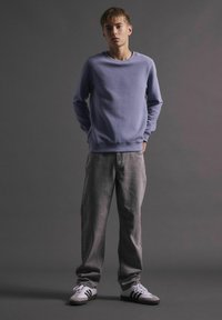 Sweatshirt mauve clair avec des poignets côtelés, associé à un pantalon gris ample et des baskets grises avec des rayures noires. Fond neutre.