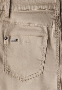 Bežová tkanina s hladkým povrchem, obsahující zadní kapsu se stříbrným knoflíkem a malou kovovou značkovou visačkou. Prošívání podél okrajů.