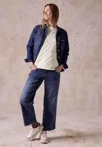 Jeansjacke, hellgrünes, gesteppte Oberteil mit geometrischem Muster, weit geschnittene blaue Jeans, weiße Sneakers mit bordeauxfarbenen Akzenten, gestrickte Socken.