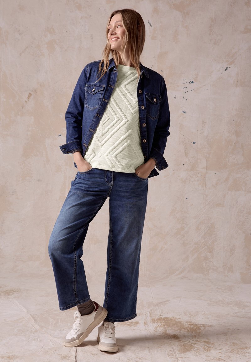 Jeansjacke, hellgrünes, gesteppte Oberteil mit geometrischem Muster, weit geschnittene blaue Jeans, weiße Sneakers mit bordeauxfarbenen Akzenten, gestrickte Socken.