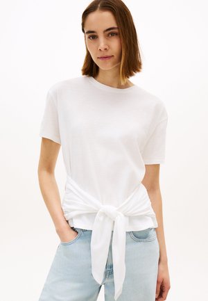 Giovane donna che indossa una maglietta bianca annodata sul davanti e jeans azzurri, in piedi con una mano in tasca davanti a uno sfondo semplice.