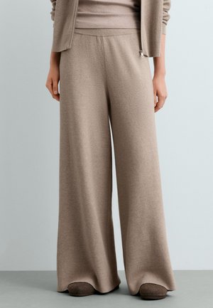 Femme portant un pantalon large en maille beige et un pull assorti avec des chaussons, debout devant un fond gris clair uni.