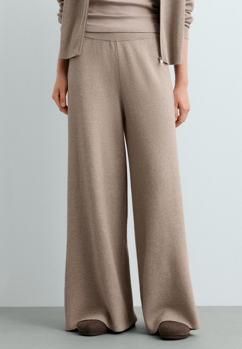 Femme portant un pantalon large en maille beige et un pull assorti avec des chaussons, debout devant un fond gris clair uni.