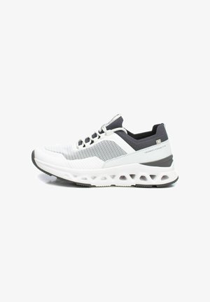 Zapatilla deportiva blanca y gris con parte superior de malla transpirable, detalles texturizados y suela acolchada con patrones recortados.