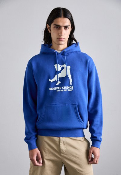 Felpa in cotone blu con tasca frontale, caratterizzata da una grafica bianca e il testo "KIDSUPER STUDIOS" sul petto. Design a pullover con coulisse.
