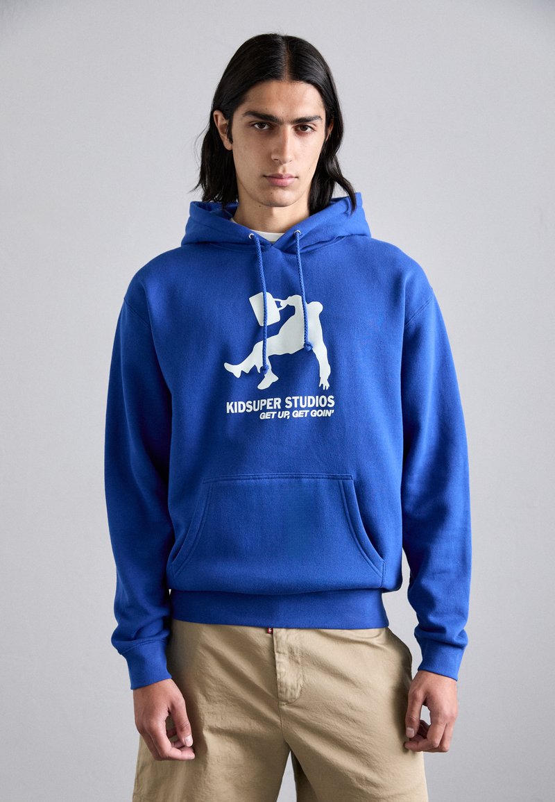 Felpa in cotone blu con tasca frontale, caratterizzata da una grafica bianca e il testo "KIDSUPER STUDIOS" sul petto. Design a pullover con coulisse.