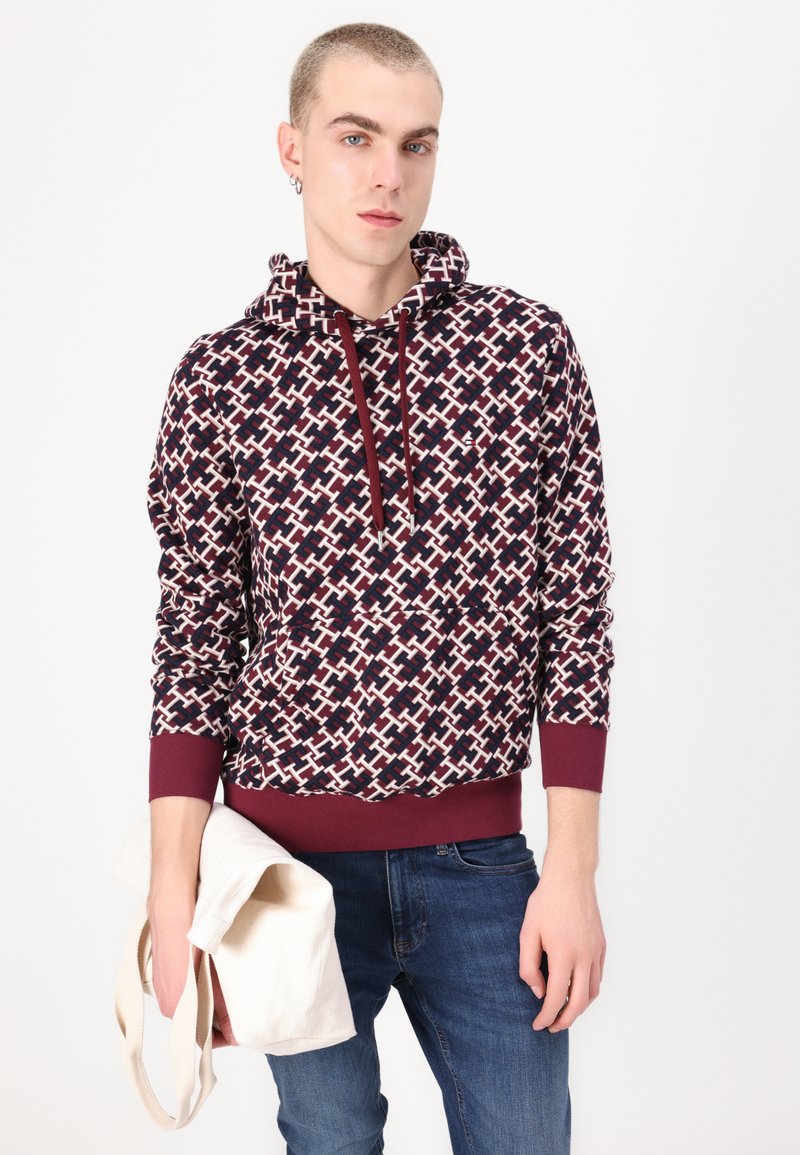 Felpa con cappuccio caratterizzata da un motivo geometrico bordeaux e crema, con polsini e orlo a costine. Il modello tiene una borsa a tracolla crema.