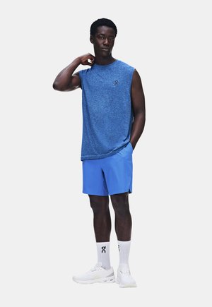 Homme portant un débardeur bleu, un short bleu, des chaussettes blanches et des baskets blanches, debout avec une main dans le cou et l’autre dans la poche.