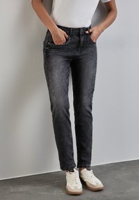 Jeans de mezclilla gris oscuro con corte recto, bolsillos frontales y cierre de botones, combinados con zapatillas blancas con acentos en beige.