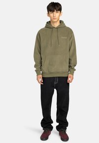 Felpa verde oliva con cappuccio, dotata di tasca frontale e lacci, abbinata a pantaloni neri e sneaker bordeaux. Logo ricamato sul petto.