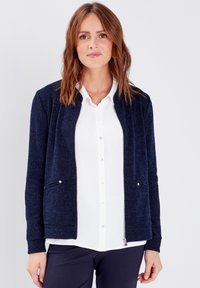 Veste bomber texturée marine avec deux poches avant, fermeture éclair et poignets côtelés, superposée sur une chemise blanche à boutons.