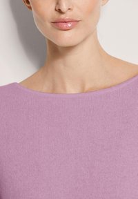Nahaufnahme des unteren Gesichts und Halses einer Person, die ein lavendelfarbenes Strickoberteil mit weitem Ausschnitt und glänzenden, nackten Lippen trägt.