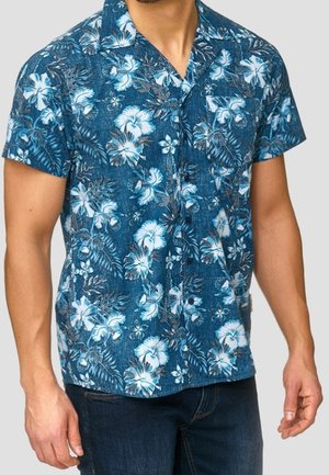 Homme portant une chemise hawaïenne bleue à manches courtes avec des motifs floraux blancs et un jean bleu foncé sur un fond uni.