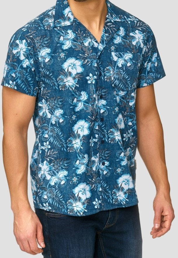 Homme portant une chemise hawaïenne bleue à manches courtes avec des motifs floraux blancs et un jean bleu foncé sur un fond uni.
