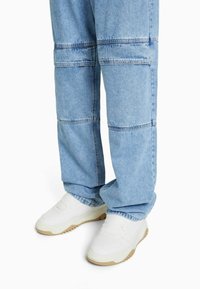 Ljusblå denim cargo-byxor kombinerade med vita sneakers med beige sulor på vit bakgrund.