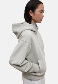 Mujer de perfil vistiendo una sudadera con cremallera de gran tamaño en gris claro, con una capucha grande y las manos en los bolsillos frontales.