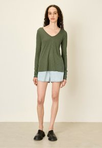 Olivno zelena dolga majica s V-izrezom, kombinirana s svetlim denim kratkimi hlačami. Model nosi črne natikače in stoji na nevtralni podlagi.
