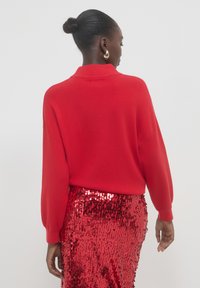 Pull en tricot rouge avec col et manches longues, associé à une jupe rouge brillante ornée de sequins. La texture est douce et les couleurs sont vives.