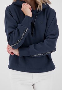Sudadera azul marina hecha de un tejido suave, con un bolsillo tipo canguro, capucha con cordón y detalles de logo en blanco en las mangas.