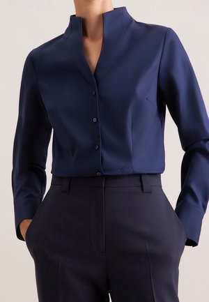 Personne portant une chemise bleu foncé boutonnée avec un col montant et un pantalon taille haute assorti bleu foncé, les mains dans les poches.