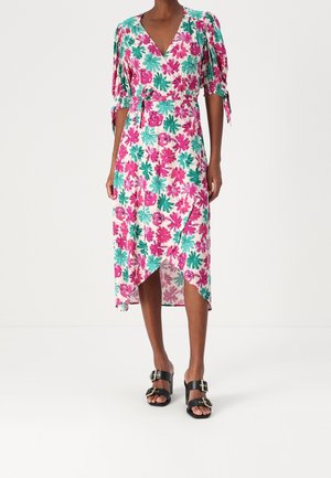 Kobieta ubrana w kwiatową sukienkę kopertową z różowo-turkusowymi kwiatami, z bufiastymi rękawami z wiązaniami oraz czarne sandały na klamrę na białym tle.