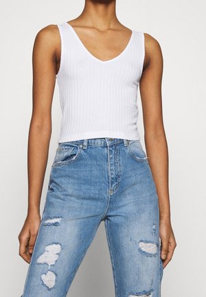 Vrouw die een wit geribd mouwloos topje met V-hals en een hoge taille lichtblauwe gescheurde jeans draagt, voor een effen achtergrond.