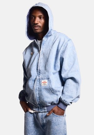 Sudadera de denim azul claro con cierre de cremallera, bolsillos frontales, puños elásticos y patrones de costura blanca detallados. Parche de logo blanco en la parte frontal.